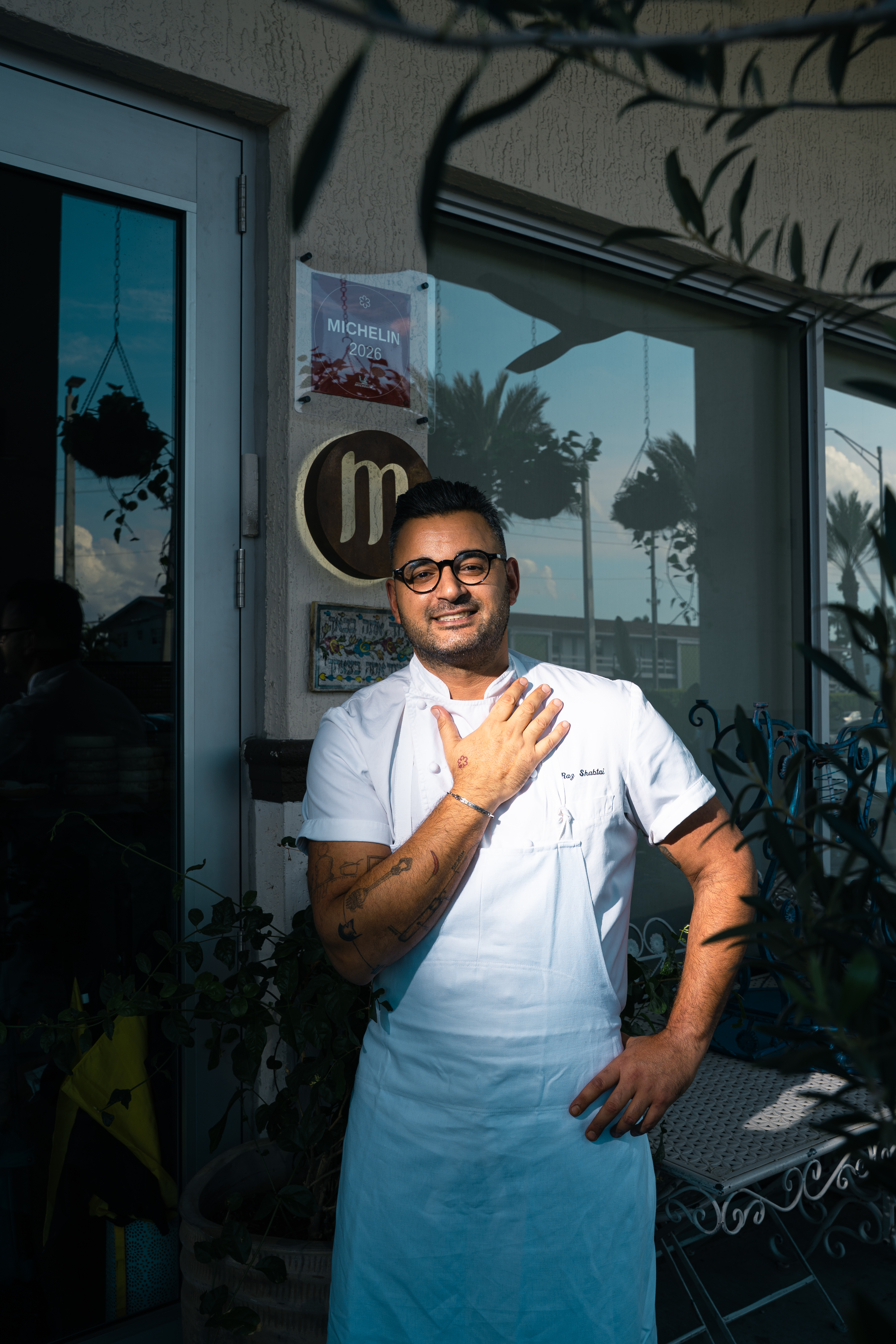 Chef portant une veste blanche et un tablier, debout devant l’entrée du restaurant Mutra.